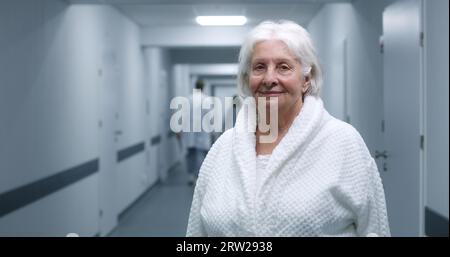 Die Seniorin steht in einem modernen Klinikkorridor. Ältere Patientin lächelt und schaut in die Kamera. Medizinisches Personal, Ärzte und Patienten im Hintergrund im Krankenhaus oder medizinischen Zentrum Flur. Hochformat. Stockfoto