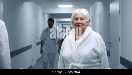 Die Seniorin steht in einem modernen Klinikkorridor. Ältere Patientin lächelt und schaut in die Kamera. Medizinisches Personal, Ärzte und Patienten im Hintergrund im Krankenhaus oder medizinischen Zentrum Flur. Hochformat. Stockfoto