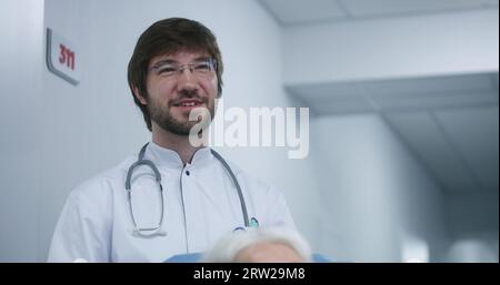 Männlicher Arzt steht im modernen Klinikkorridor. Der Facharzt kommuniziert mit der Kollegin oder dem Patienten, diskutiert die Behandlung oder Testergebnisse. Medizinisches Personal und Patienten im Spitalgang. Stockfoto