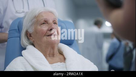 Ältere Frau im Transferrollstuhl kommuniziert mit dem Arzt im Klinikkorridor. Die Patientin spricht vor der Therapie mit der Ärztin. Medizinisches Personal und Patienten, die im Hintergrund im Krankenhausflur gehen. Stockfoto