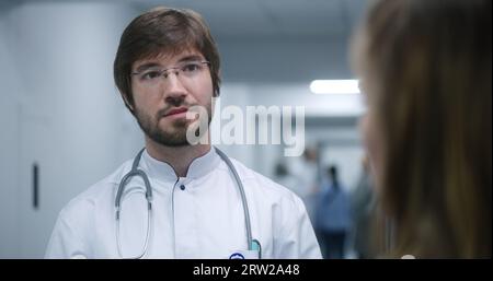 Männlicher Arzt steht im modernen Klinikkorridor. Der Facharzt kommuniziert mit der Kollegin oder dem Patienten, diskutiert die Behandlung oder Testergebnisse. Medizinisches Personal und Patienten im Spitalgang. Stockfoto