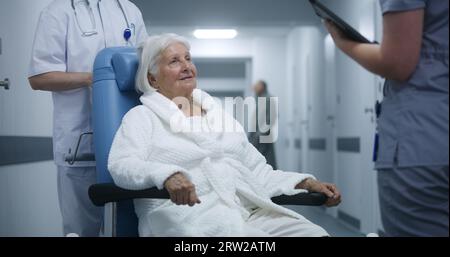 Ältere Frau im Rollstuhl spricht mit dem Arzt im Klinikkorridor. Medic diskutiert Testergebnisse mit dem Patienten, verwendet digitales Tablet. Medizinisches Personal und Patienten laufen im Hintergrund im Krankenhausflur. Stockfoto