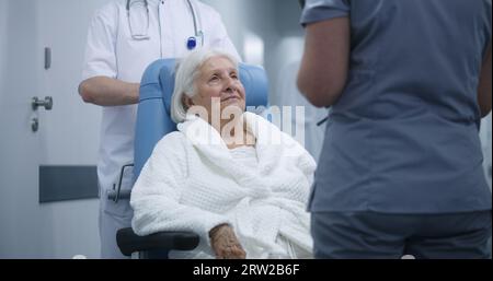 Ältere Frau im Rollstuhl kommuniziert mit dem Arzt im Klinikkorridor. Die Patientin spricht mit der Ärztin vor Eingriffen oder Therapien. Medizinisches Personal und Patienten im Hintergrund im Spitalgang. Stockfoto
