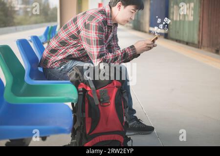 Junger asiatischer Rucksackreisender mit rotem kariertem Hemd sitzend und Nutzer-Smartphone, um E-Mails zu lesen, Nachrichten mit Freunden am Bahnsteig auszutauschen Stockfoto