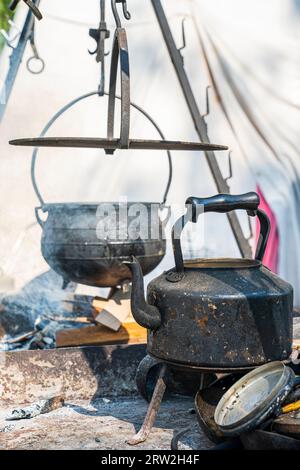 Mittelalterlicher schwarzer Wasserkocher aus Eisen auf einem gusseisernen Ständer, dahinter ein unscharfer Kessel, der über einem schwelenden Feuer hängt. Mittelalterliches Sandwich-Zentrum. Stockfoto