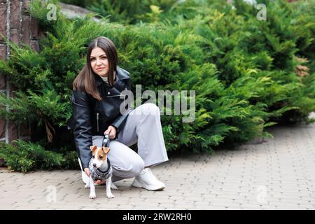 Frau in einer legeren Jacke, die am Herbsttag in der Nähe ihres Lieblingshundes sitzt, der in einem Anzug für Hunde auf der Straße trainiert ist Stockfoto