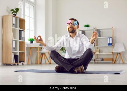 Lustiger Mann mit Haftnotizen im Gesicht, der im Büro auf dem Boden sitzt, Meditation macht und sich entspannt Stockfoto