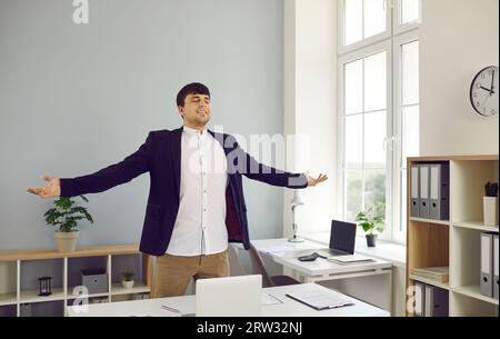 Junger Büroangestellter steht neben dem Schreibtisch mit Geschäftsdokumenten und spreizt die Arme zur Seite Stockfoto