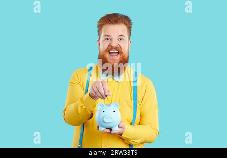 Lustiger rothaariger Kaukasier mit Bart und Schnurrbart wirft Münzen in die Sparkasse, um Geld zu sparen Stockfoto