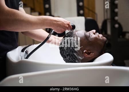 Der Mann wäscht Schaum von den Haaren des Kunden mit warmem Wasser ab Stockfoto