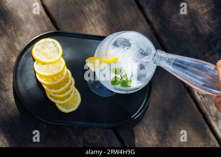 Draufsicht auf eine Flasche Tonic, die über ein Glas mit Eis gegossen wurde, eine Scheibe Zitronenblätter und Minzblätter, Zubereitung eines Gins und Tonic Stockfoto