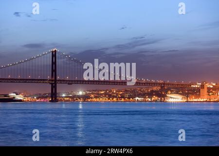 25. April (Portugiesisch: Ponte 25 de Abril) Brücke über den Tejo bei Nacht und Stadt Lissabon, Portugal im Hintergrund Stockfoto