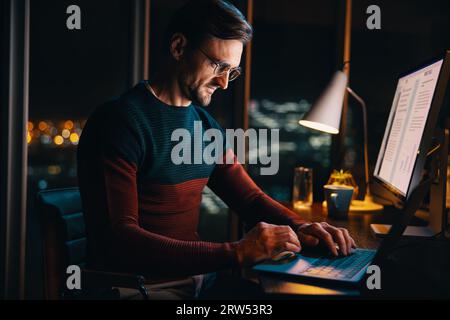 Ein Geschäftsmann, der spät von zu Hause aus arbeitet und auf einem Laptop schreibt. Engagiert und motiviert setzt er Prioritäten für sein Projekt und engagiert sich für Remote-Arbeit. Stockfoto