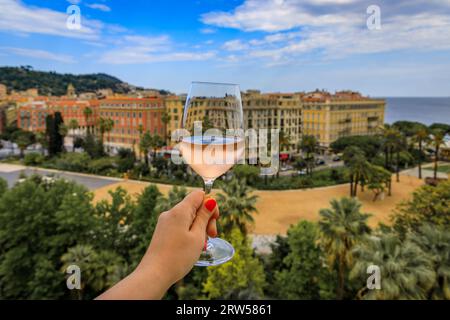 Die Hand der Frau hält ein Glas provenzalischen Roséweins in einem Restaurant im Freien mit einem Hintergrund verschwommener Gebäude in der Altstadt von Nizza, Südfrankreich Stockfoto