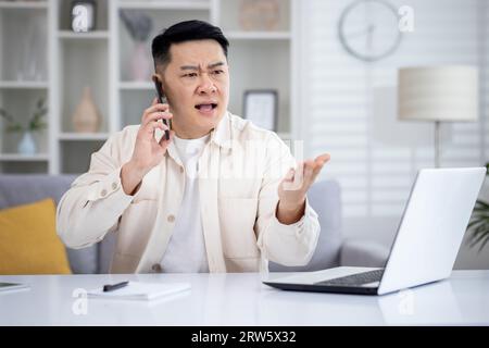 Wütender, wütender und verärgerter Mann, der zu Hause am Telefon spricht, asiate, die aus der Ferne vom Home Office aus arbeiten, am Tisch im Wohnzimmer sitzen, freischaffender Geschäftsmann bei der Arbeit. Stockfoto