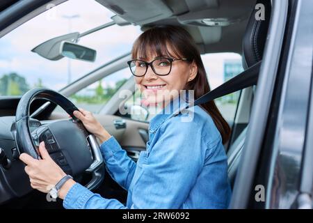 Eine Fahrerin mittleren Alters sitzt im Auto hinter dem Steuer und schaut lächelnd auf die Kamera Stockfoto