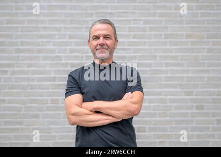 Vorderansicht eines attraktiven Mannes mit gekreuzten Armen, der freundlich und entschlossen auf die Kamera vor einer weißen Backsteinmauer schaut Stockfoto