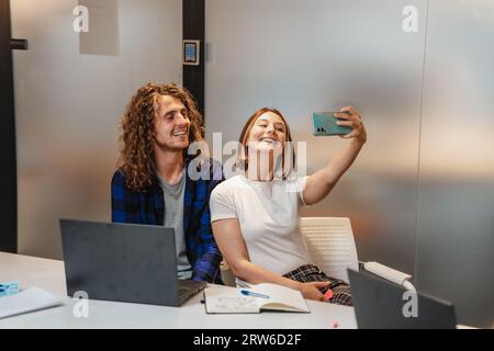 Seitenansicht eines Paares, das ein Selfie bei der Arbeit macht. Kurzhaariges Mädchen, das ein Selfie mit seiner Kollegin macht, während es eine Pause bei der Arbeit macht Stockfoto