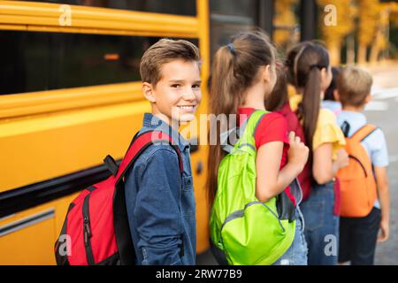 Porträt Eines Lächelnden Jungen Mit Rucksack, Der Mit Klassenkameraden In Den Gelben Schulbus Einsteigt Stockfoto