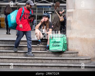 Uber isst Fahrradkurierfahrerin, die etwas Essen isst, während sie im Zentrum Londons auf Bestellungen wartet, wobei Deliveroo-Fahrer vorbeiläuft Stockfoto