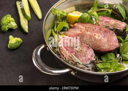 Geschnittener Flussfisch mit Minze und Rosmarin in der Pfanne. Brokkoli und Gurkenscheiben auf dem Tisch. Schwarzer Hintergrund. Draufsicht Stockfoto