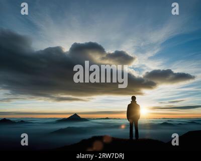 Silhouette eines Mannes, der einen friedlichen Sonnenaufgang in den Bergen genießt, Nebel, der um die Gipfel lauert, Sonne, die zwischen den Wolken brennt Stockfoto