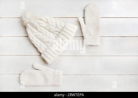 Eine weiße Strickmütze mit Pompon und Wollhandschuhen liegt auf einem weißen hölzernen Hintergrund. Details der Winterkleidung, flach liegend, Draufsicht Stockfoto