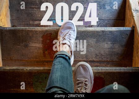 Frauenbeine auf einer Holztreppe und der Text 2024 auf der Treppe, gehen Sie die Treppe hinunter, frohes neues Jahr 2024, los Stockfoto