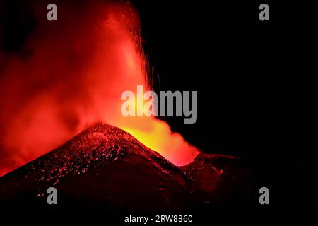 Ätna - massive, geschmolzene Lava, die im Detail von der Silhouette des Vulkankraters im dunklen Nachthintergrund und den starken Rauch- und Dampfemissionen sprüht Stockfoto