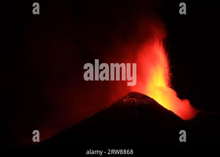 Ätna - massive, geschmolzene Lava, die im Detail von der Silhouette des Vulkankraters im dunklen Nachthintergrund und den starken Rauch- und Dampfemissionen sprüht Stockfoto