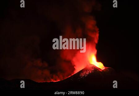 L'Etna in un'eruzione lavica notturna con cratere sagomato in controluce e grandi emissioni di fumo in una vista panoramica del vulcano siciliano Stockfoto