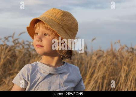 Ein kleiner Junge in einem gelben Panamahut steht vor dem Hintergrund eines landwirtschaftlichen Feldes in den Strahlen der untergehenden Sonne und schaut nach links Stockfoto