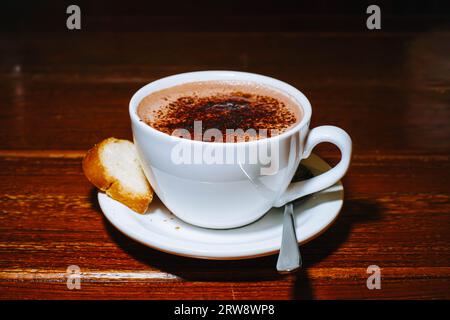 Eine Tasse Cappuccino mit einem Brot in einer weißen Porzellan-Achterbahn und einem Löffel auf einem Holztisch Stockfoto