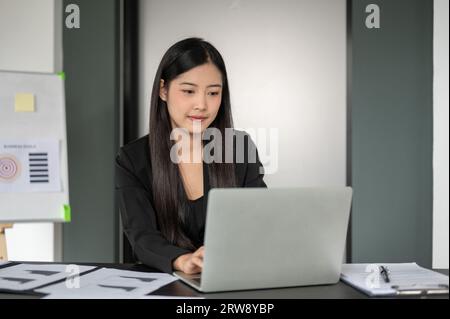 Eine schöne asiatische Geschäftsfrau in einem formellen schwarzen Anzug konzentriert sich auf ihr Unternehmensprojekt auf ihrem Laptop und analysiert geschäftliche Finanzdaten auf ihrem com Stockfoto
