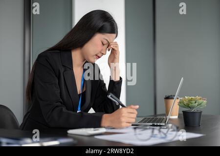Eine gestresste und nachdenkliche asiatische Geschäftsfrau oder Buchhalterin, die an ihrem Schreibtisch im Büro arbeitet, unter Kopfschmerzen leidet und sich Sorgen um abo macht Stockfoto