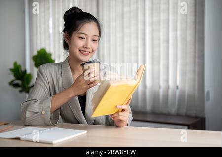 Eine schöne und glückliche asiatische Geschäftsfrau in einem lässigen Anzug liest einige Details auf ihrem Notebook, während sie in ihrer Mo einen Morgenkaffee an ihrem Schreibtisch trinkt Stockfoto