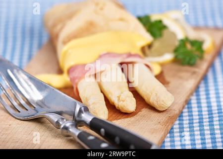 Weißer Spargel mit Schinken und Omelette liegt auf einem rustikalen Holzbrett. Rustikales Besteck liegt links vorne. Das Bild hat ein feines Bokeh Stockfoto