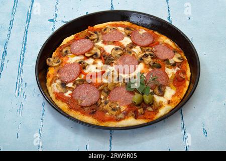 Frisch gebackene Pizza in einer Pizzaform, garniert mit Salami, Tomaten, Käse, Oliven, Pilz, Zwiebeln, garniert mit zwei Oliven und einem Haufen Rosmarin und Stockfoto