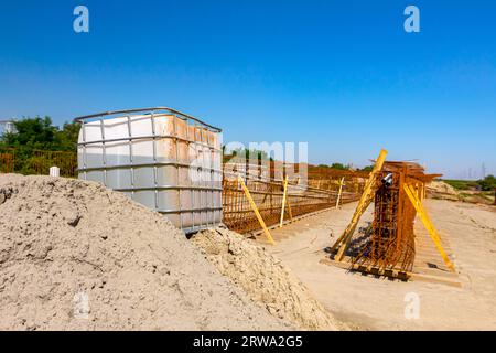 Weißer quadratischer Wassertank, kubisch, mit Metallgitter, riesiger Flüssigkeitsbehälter auf Holzpaletten auf der Baustelle, im Hintergrund ist Brückengerüst-Reinforc Stockfoto