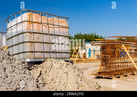 Weißer quadratischer Wassertank, kubisch, mit Metallgitter, riesiger Flüssigkeitsbehälter auf Holzpaletten auf der Baustelle, im Hintergrund ist Brückengerüst-Reinforc Stockfoto