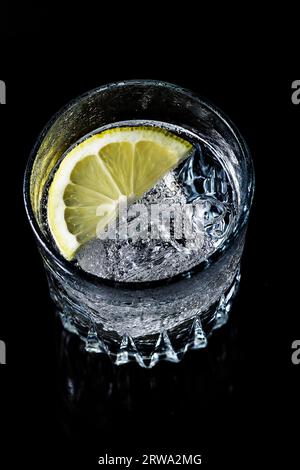 Ein Glas Mineralwasser, das von oben auf einem schwarzen Hintergrund fotografiert wurde. Das Glas enthält Wasser, Eiswürfel und eine halbe Zitronenscheibe. Ein Glas Stockfoto