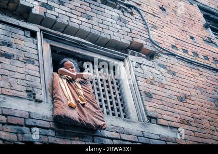 Bhaktapur, Nepal, um Juni 2012: Ein junges Mädchen schaut aus einem Holzfenster in einem Backsteinhaus in Bhaktapur, Nepal. Dokumentarisches Editorial Stockfoto