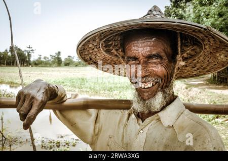 Srimongal, Bangladesch, um Juli 2012: Der alte Mann mit grauem Bart und großem Strohhut hält in Srimongal, Bangladesch, einen Holzstock auf der Schulter. Stockfoto