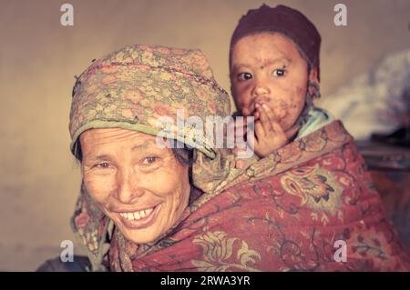 Dolpo, Nepal, um Mai 2012: Foto einer gebürtigen braunen Frau im Kopftuch, die ein Kind mit brauner Kappe auf dem Rücken trägt, und in Dolpo, Nepal. Stockfoto