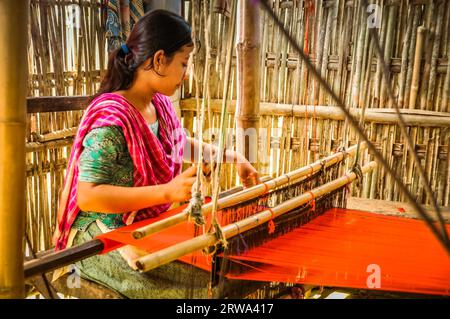 Srimongal, Bangladesch, um Juli 2012: Junge schwarzhaarige Frau mit rosa Schal sitzt im Unterschlupf und webt auf einfachen Holzwebstuhl in Srimongal Stockfoto