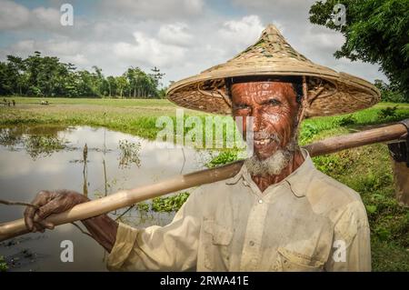 Srimongal, Bangladesch, um Juli 2012: Ein alter Mann mit Falten im Gesicht und grauem Bart hält in Srimongal einen Holzstock auf der Schulter Stockfoto