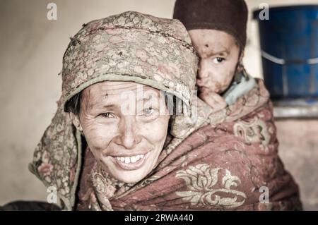 Dolpo, Nepal, um Mai 2012: Die gebürtige Frau trägt ein Kind mit brauner Kappe auf dem Rücken in Dolpo, Nepal. Dokumentarisches Editorial Stockfoto