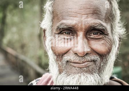 Sunderban, Bangladesch, um den Juli 2012: Alter Einheimischer mit weißem Bart und Haaren trägt einen farbenfrohen Schal und ein braunes Hemd und hält einen Holzstab in der Hand Stockfoto