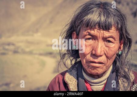 Dolpo, Nepal, um Juni 2012: Alte grauhaarige Frau mit faltigem Gesicht trägt blaue Ohrringe und sieht traurig aus, als ob sie in Dolpo, Nepal, Fotokamera wäre. Stockfoto
