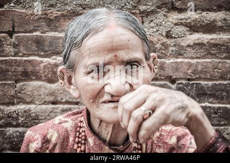 Bhaktapur, Nepal, um Juni 2012: Alte grauhaarige Frau mit faltigem Gesicht in roten Kleidern mit Kette aus Perlen trägt Ohrringe in Stockfoto